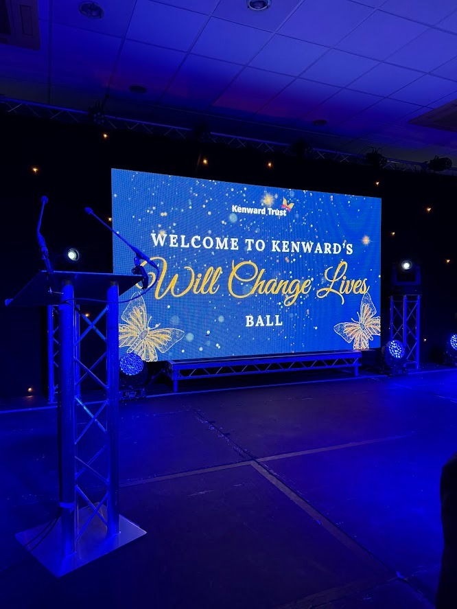 The stage screen at the Kenward Trust ball reading “Welcome to Kenward’s Will Change Lives Ball”